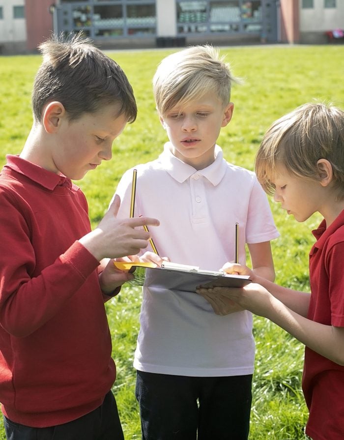 three boys gathered around a clipboard for an outdoor lesson on the school field