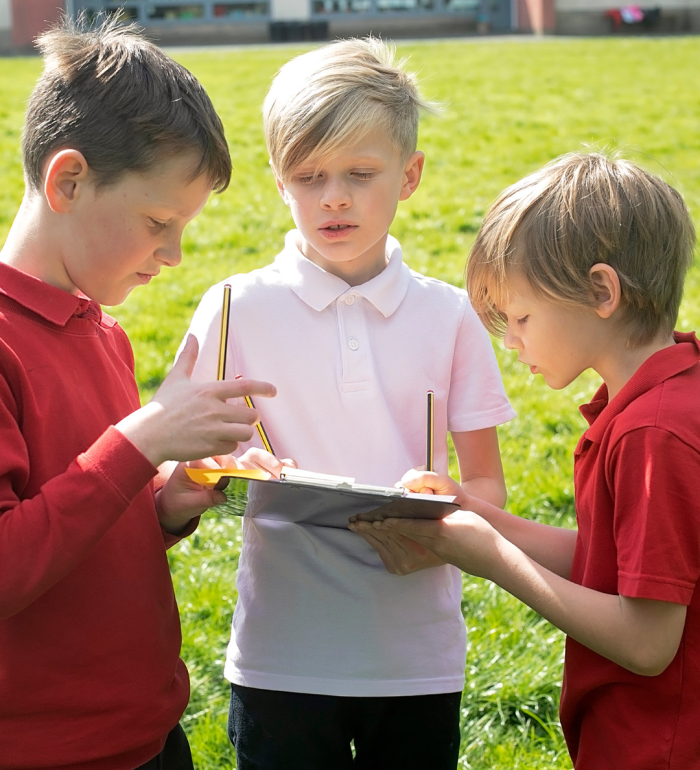 3 boys working together on a clipboard to solve a problem on the school field
