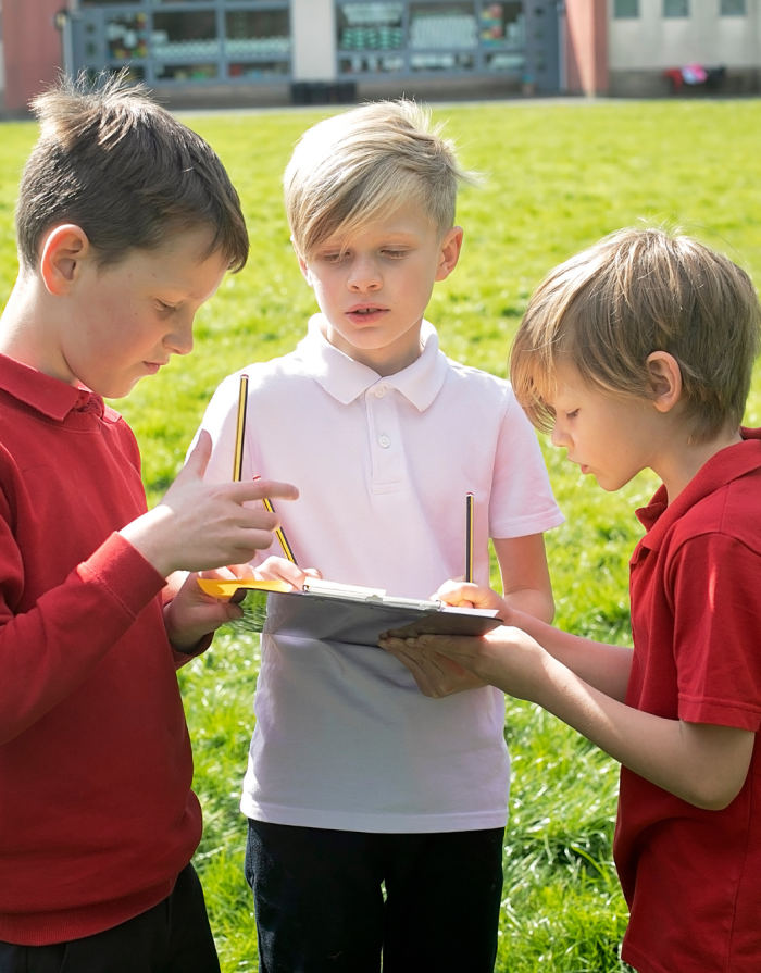3 boys working together on a clipboard to solve a problem on the school field