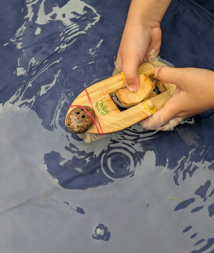 a child winding up a wooden paddle boat with a pebble person attached