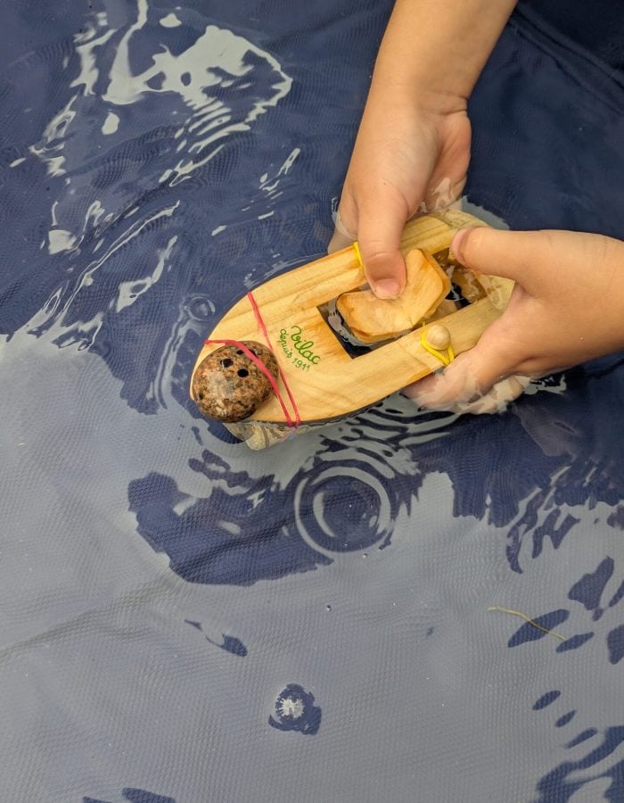 a child winding up a wooden paddle boat with a pebble person attached
